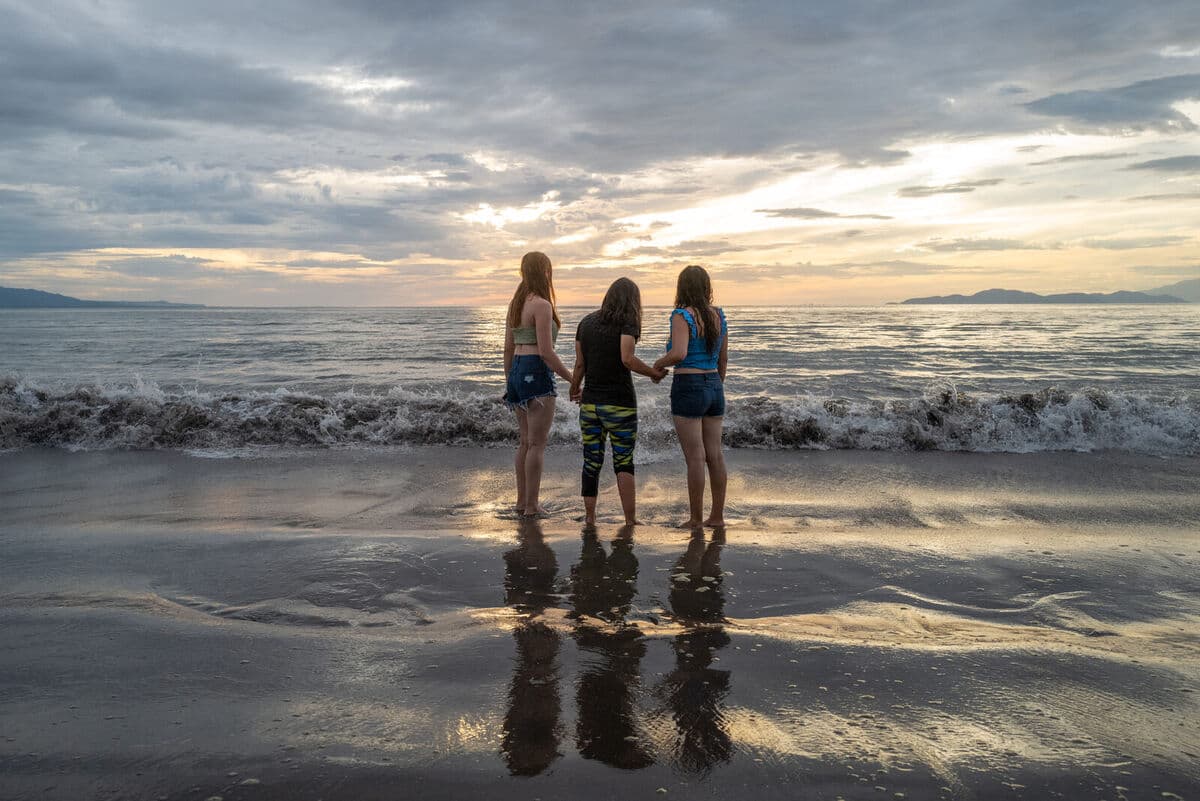 Three people holding hands facing the sea at sunset